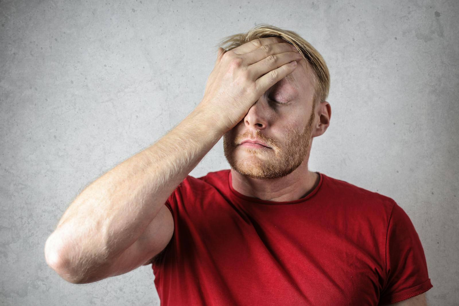Homem corta o bingulim para cumprir aposta A frustrated man in a red shirt holds his head in stress against a neutral background.
