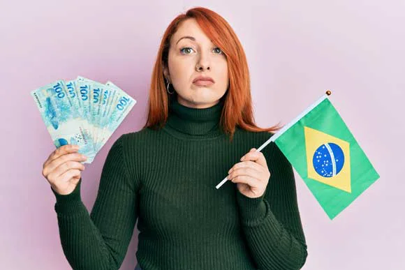 hermosa mujer pelirroja sosteniendo 100 billetes reales brasilenos y bandera de brasil relajada con expresion seria en la cara mirada simple y natural la camara | Dicas | dicas